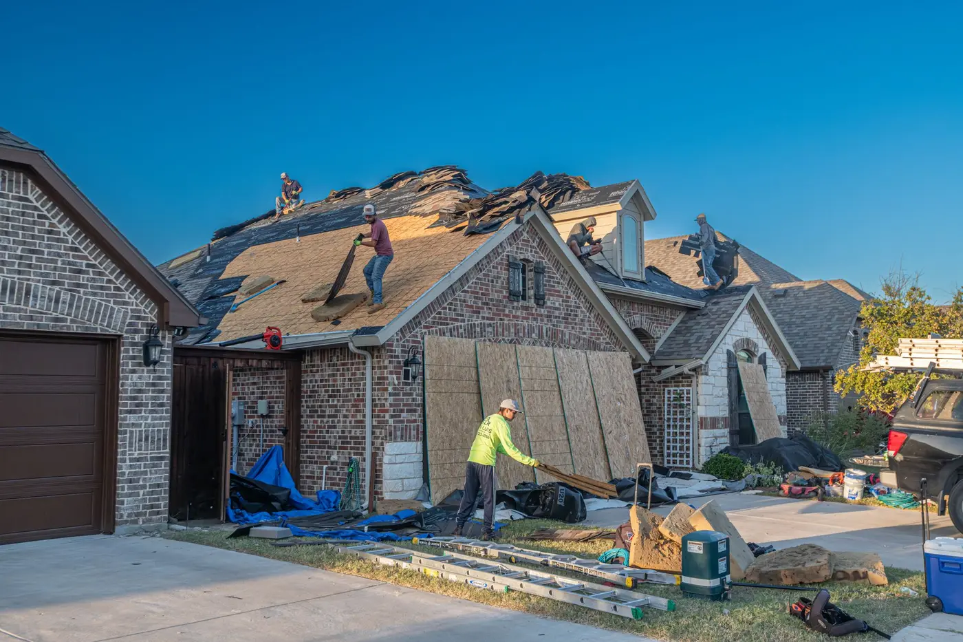 A crew stripping and re-decking the roof of a brick house, with tear-off debris tarped on the driveway below
