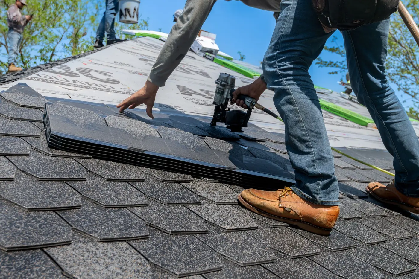 A roofer kneeling on a steep roof, setting a bundle of architectural shingles above a line of synthetic underlayment