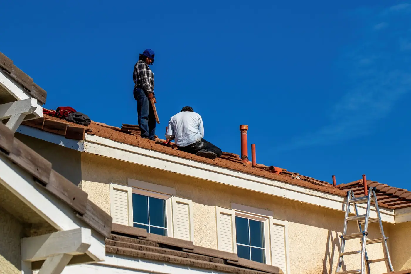 Two roofers working a small section of tile at the eave of a house, a ladder set against the gutter beside them