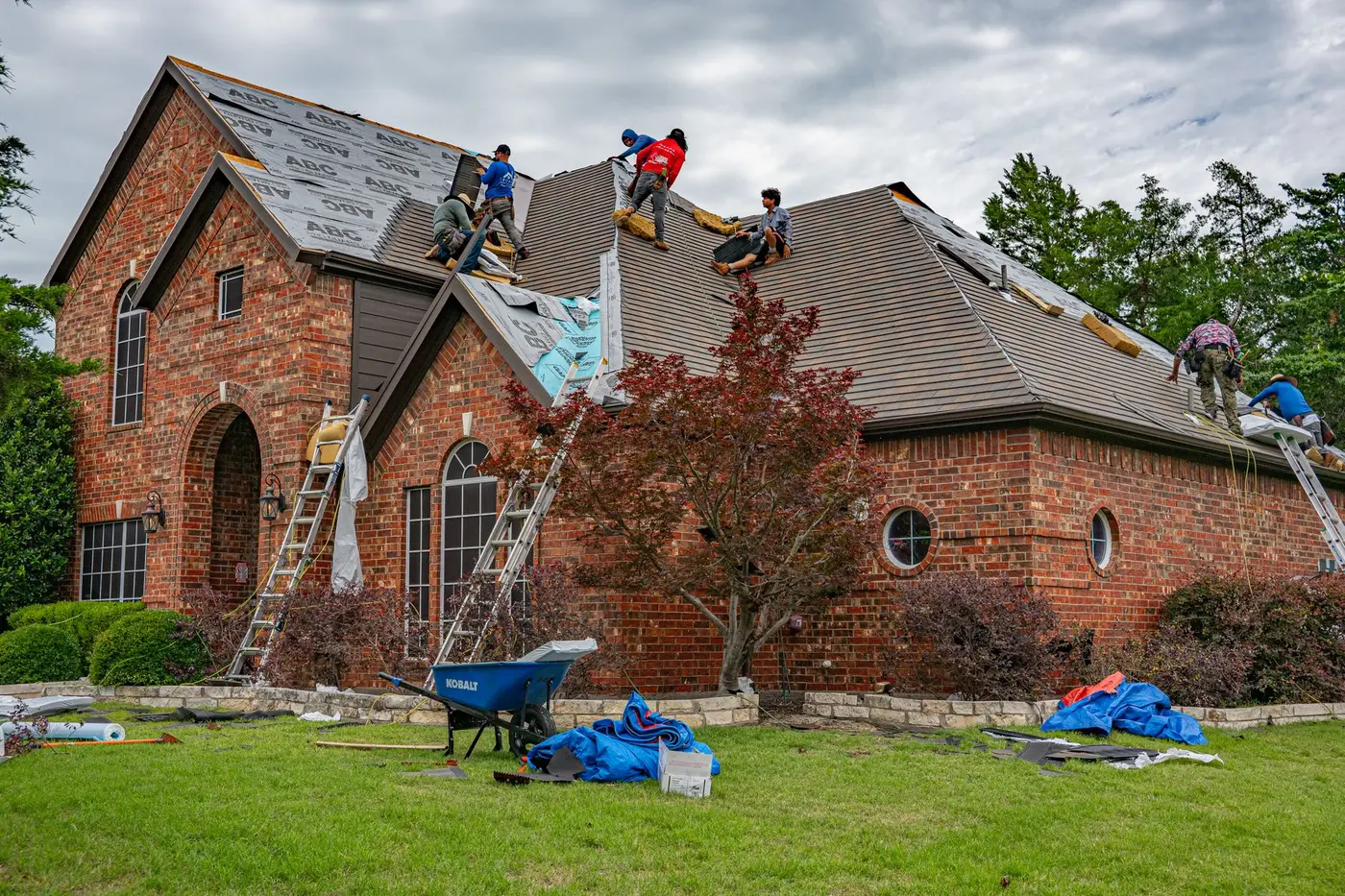 A six-person crew working across the roof of a brick house, ladders up on two elevations and tarps laid out on the lawn