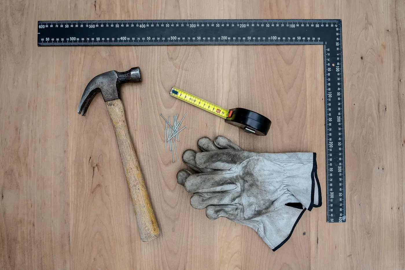 A framing square, claw hammer, tape measure, nails and leather work gloves laid out on a wooden board